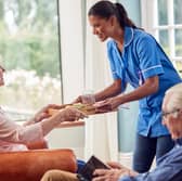 Female Care Worker In Uniform Bringing Meal On Tray To Senior Woman Sitting In Lounge At Home