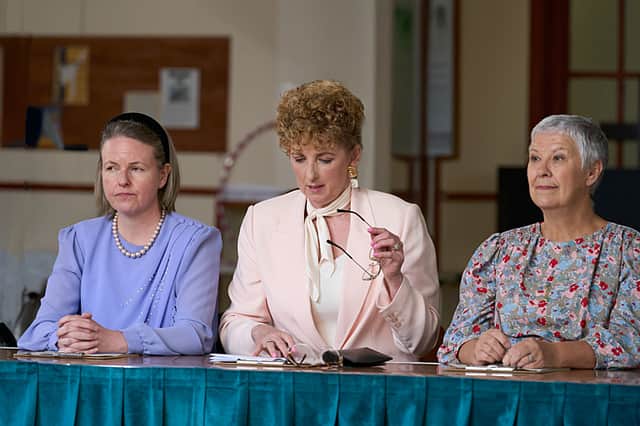 Three judges for the Govan Fair Queen contest seated in a row in a town hall. The head judge, played by Julie Wilson Nimmo, sits in the middle