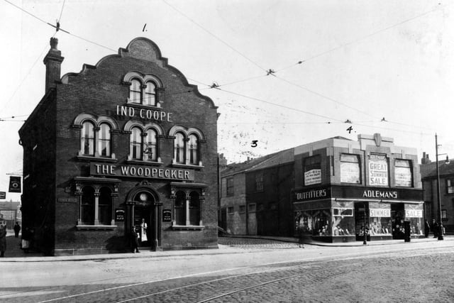 The Woodpecker Inn located at Number 1 York Road, with Burmantofts Street to the left and Waterloo Place to the right in 1935. There are two men in the doorway. A tram fare stage sign is on the left hand side of the pub and there are tram lines in the road. On the right is Adleman's Clothier's located on York Road, which advertises a summer sale