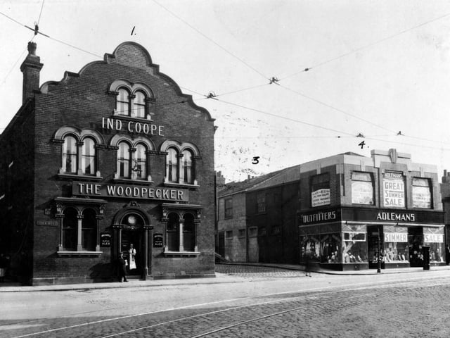 The Woodpecker Inn located at Number 1 York Road, with Burmantofts Street to the left and Waterloo Place to the right in 1935. There are two men in the doorway. A tram fare stage sign is on the left hand side of the pub and there are tram lines in the road. On the right is Adleman's Clothier's located on York Road, which advertises a summer sale