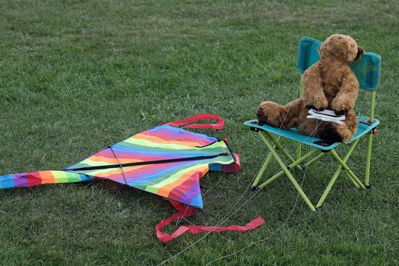 A teddy bear takes a break from flying a rainbow kite.