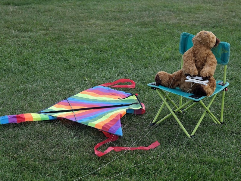 A teddy bear takes a break from flying a rainbow kite.