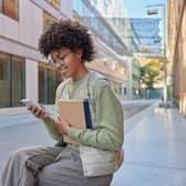 Glad student has curly hair holds notebook uses mobile phone for surfing internet reads ideas for project dressed casually poses outdoor.