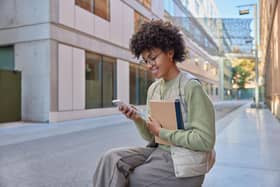 Glad student has curly hair holds notebook uses mobile phone for surfing internet reads ideas for project dressed casually poses outdoor.