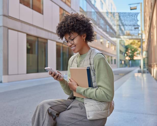 Glad student has curly hair holds notebook uses mobile phone for surfing internet reads ideas for project dressed casually poses outdoor.