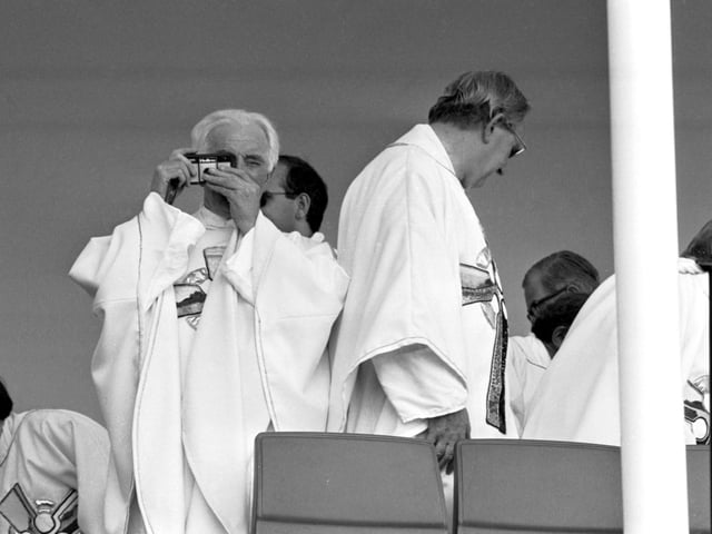 An unidentified priest uses his camera, with his finger over the lens, to take pictures of Pope John Paul II at Bellahouston Park.