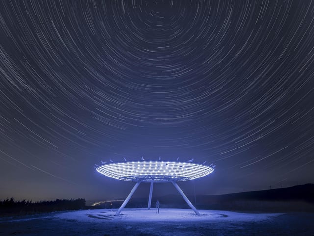 Highly Commended
Close Encounters of The Haslingden Kind © Katie McGuinness
Haslingden’s Halo is an 18-meter diameter sculpture located in the hills of Lancashire. McGuinness took inspiration from the Close Encounters of The Third Kind film poster to create her image. More than 150 images, taken over an hour and with exposures of 25 seconds each, were combined to show the apparent rotation of stars around Polaris.
‘Positioned just above the town of Haslingden is a sculpture, one of four designed to encourage people to walk in the countryside and enjoy the marvels of Lancashire. This sculpture is known as the Halo and it is illuminated by hundreds of LED lights. The photographer has been very creative taking over 150 images in an hour, which were then merged in Photoshop to produce this masterpiece. The category of People and Space is a marvellous opportunity for talented photographers to get creative and this is an example of what can be achieved by being aware of your surroundings and being alert to the potential for award-winning photography.’ - Alan Sparrow
Taken with a Sony ILCE-7M4 camera, 20 mm f/4, ISO 160; Sky: 151 x 25-second exposures; Foreground: 5-second exposure
Location: Haslingden, Lancashire, UK