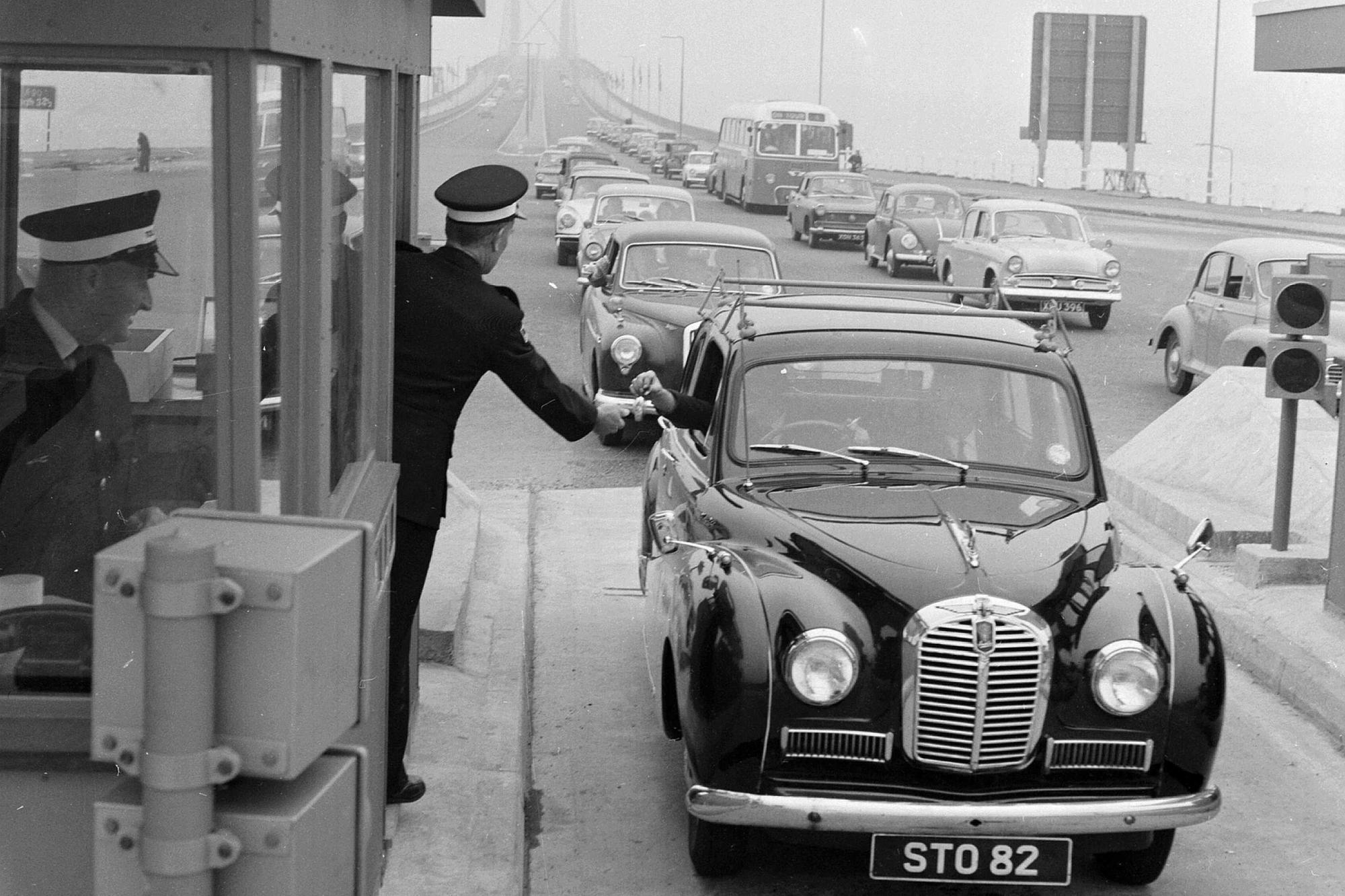 A driver pays the toll at the Forth Road Bridge in 1964, handing the money to a uniformed collector ... and how cars have changed too!