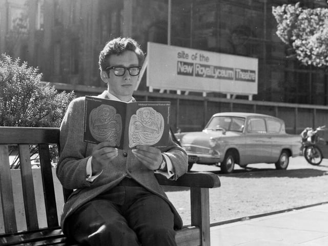 A visitor sits in Castle Terrace reading the Edinburgh International Festival guide book in August 1966. The sign behind him reads 'Site of the New Royal Lyceum Theatre'.