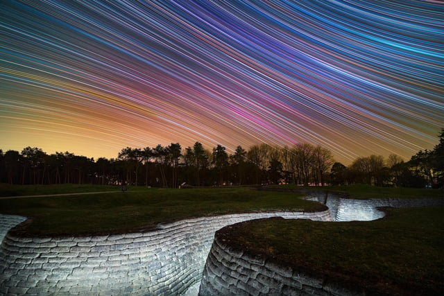 Runner-Up
Celestial Equator Above First World War Trench Memorial © Louis Leroux-Gere
Star trails above the preserved First World War trenches in Canadian National Vimy Memorial Park, Northern France. Taken over five hours, the camera captured the rotation of the sky, revealing the colourful stars.  
‘In April 1917, Canadian and German troops fought and died on this ridge near Vimy in northern France. The Canadian Government later preserved the cratered battlefield as a war memorial. This haunting image contrasts the conflict-scarred landscape with the ethereal beauty of the stars. A line of trees divides the composition into two distinct halves. Above, pastel-coloured star trails trace smooth arcs across the sky. Below, a trench carves a twisted fissure through the dull green grassland. The result is a potent reminder of how human wars have disfigured the surface of our planet.’ – Katherine Gazzard
Taken with a Canon EOS 6D (Astro modified), Samyang XP 14 mm f/2.4 lens, 14 mm f/3.2, ISO 1000, 577 x 30-second exposures

Location: Vimy, Pas-de-Calais, Hauts de France, France