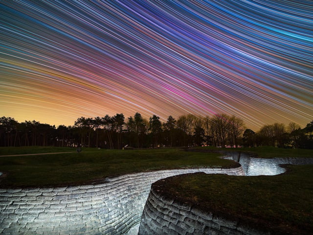 Runner-Up
Celestial Equator Above First World War Trench Memorial © Louis Leroux-Gere
Star trails above the preserved First World War trenches in Canadian National Vimy Memorial Park, Northern France. Taken over five hours, the camera captured the rotation of the sky, revealing the colourful stars.
‘In April 1917, Canadian and German troops fought and died on this ridge near Vimy in northern France. The Canadian Government later preserved the cratered battlefield as a war memorial. This haunting image contrasts the conflict-scarred landscape with the ethereal beauty of the stars. A line of trees divides the composition into two distinct halves. Above, pastel-coloured star trails trace smooth arcs across the sky. Below, a trench carves a twisted fissure through the dull green grassland. The result is a potent reminder of how human wars have disfigured the surface of our planet.’ – Katherine Gazzard
Taken with a Canon EOS 6D (Astro modified), Samyang XP 14 mm f/2.4 lens, 14 mm f/3.2, ISO 1000, 577 x 30-second exposures
Location: Vimy, Pas-de-Calais, Hauts de France, France