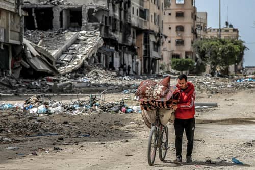 A man walks with a bicycle loaded with blankets and cushions past destroyed buildings along a street in Gaza City (Photo by -/AFP via Getty Images)