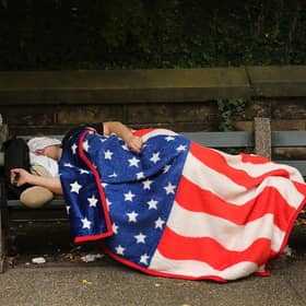 A homeless man sleeps under an American flag blanket on a park bench in Brooklyn, New York City (Picture: Spencer Platt/Getty Images)