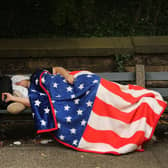 A homeless man sleeps under an American flag blanket on a park bench in Brooklyn, New York City (Picture: Spencer Platt/Getty Images)