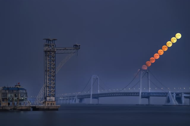 Highly Commended
Moon at Nightfall © Haohan Sun
A photograph of a moonrise over the Xinghai Bay Bridge in Dalian. Atmospheric extinction alters the hue and brightness of the Moon when it is low on the horizon. In this photo, you can see the Moon appears brighter and less red as it rises in the sky.
‘The composition of this photograph is superb, capturing the striking contrast between urban construction and the ineffable mystery of the natural world: the imposing mist, the moody sea and the motion of the Moon. While the bridge spans the horizon, the Moon also seems to stand as a bridge, or perhaps stepping stones, into the sky. I particularly liked the progression of colours on the rising Moon, from a molten red to a more familiar rocky silver, reminding us of the profound connection between human industry and the cosmos, and yet the vast expanse that separates them. A great shot from our young astronomy photographer!’ - Imad Ahmed	

Moon: Canon 24-105mm camera, 5 x ISO 1600, 6 x SO3200, 2 x 1/100-second exposures, 3 x 1/50-second exposures, 6 x 1/30-second exposures; Foreground: Canon EOS 6D Mark II camera, 150 mm f/4, ISO 1600, 40 x 1/125-second exposures

Location: Dalian, Liaoning, China