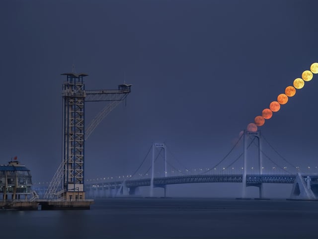 Highly Commended
Moon at Nightfall © Haohan Sun
A photograph of a moonrise over the Xinghai Bay Bridge in Dalian. Atmospheric extinction alters the hue and brightness of the Moon when it is low on the horizon. In this photo, you can see the Moon appears brighter and less red as it rises in the sky.
‘The composition of this photograph is superb, capturing the striking contrast between urban construction and the ineffable mystery of the natural world: the imposing mist, the moody sea and the motion of the Moon. While the bridge spans the horizon, the Moon also seems to stand as a bridge, or perhaps stepping stones, into the sky. I particularly liked the progression of colours on the rising Moon, from a molten red to a more familiar rocky silver, reminding us of the profound connection between human industry and the cosmos, and yet the vast expanse that separates them. A great shot from our young astronomy photographer!’ - Imad Ahmed
Moon: Canon 24-105mm camera, 5 x ISO 1600, 6 x SO3200, 2 x 1/100-second exposures, 3 x 1/50-second exposures, 6 x 1/30-second exposures; Foreground: Canon EOS 6D Mark II camera, 150 mm f/4, ISO 1600, 40 x 1/125-second exposures
Location: Dalian, Liaoning, China