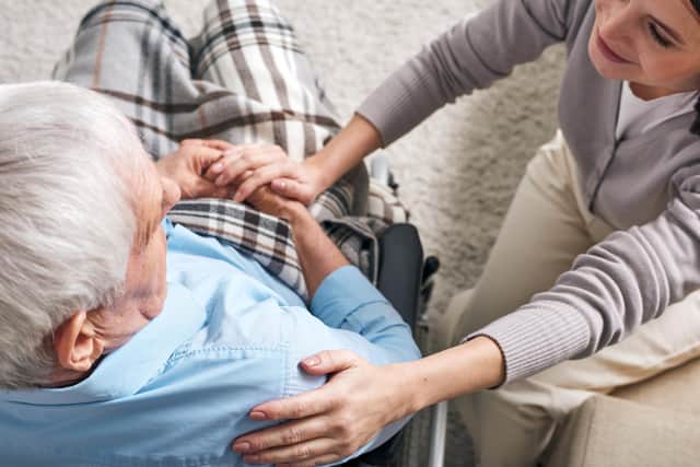 Young supportive female caregiver sitting by senior man in wheelchair and keeping her hand on his shoulder while comforting him