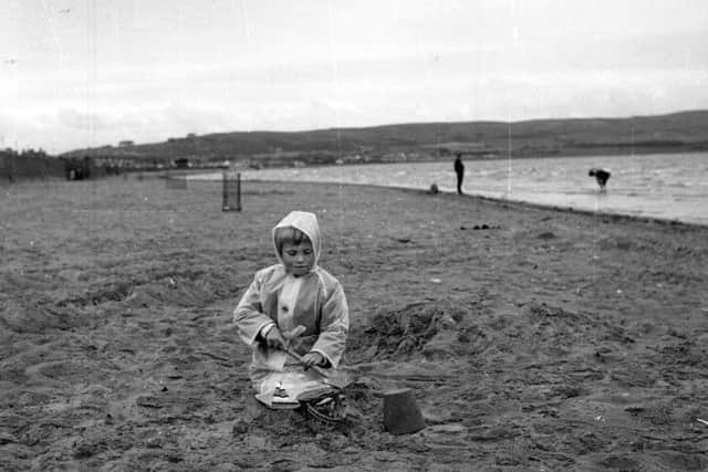 Wrapped up in her plastic Mac, Lynn McPherson makes sand pies in the summer rain on the beach at Ayr, July 1965. Picture: TSPL