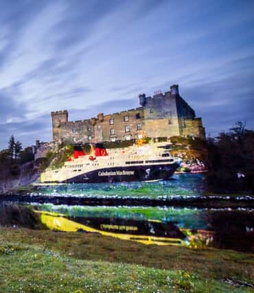An image of a Caledonian Macbrayne Ferry is projected onto Dunvegan Castle. The castle is sitting on the side of a loch and the image of the castle and thr projection are reflected in the water of the loch. Overhead there are clouds moving across the sky