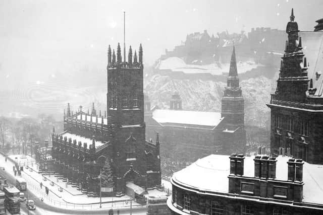 Snow falls on the  West End of Princes Street with St John's church and St Cuthbert's church in the foreground (and a Christmas tree to left of the entrance) and the frontage of the Caledonian Hotel to the right.